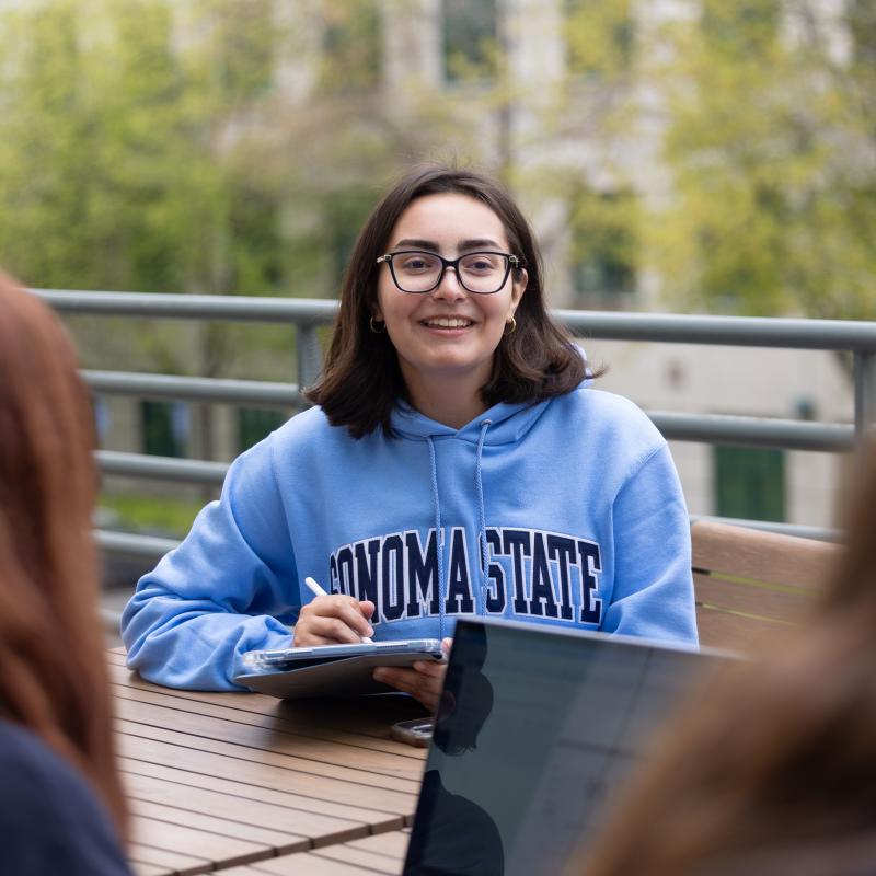 Student wearing a sweatshirt and sitting outside writing on an ipad and looking towards other students
