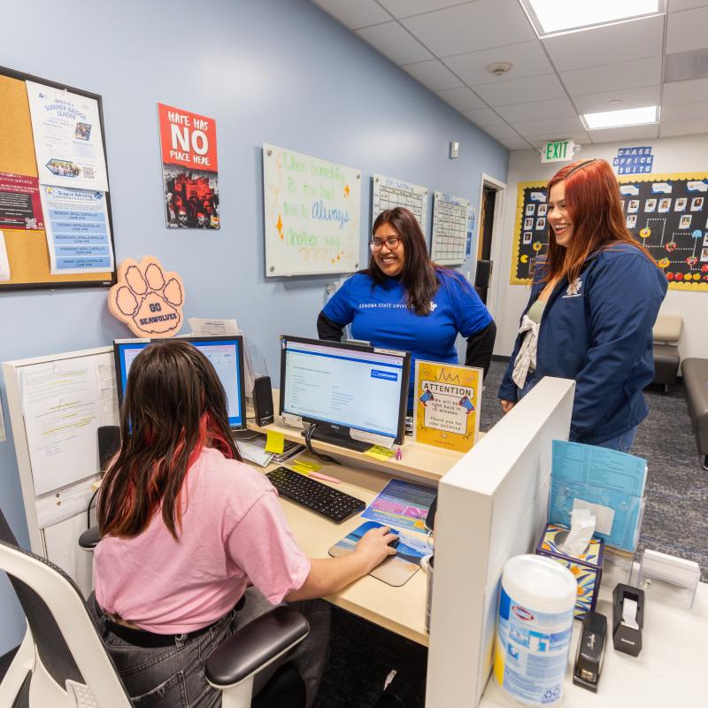 Three students at a check in desk with one looking at a computer