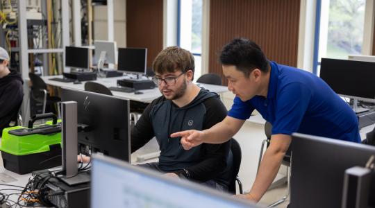 Professor standing and pointing to a computer screen with student sitting infront of computer