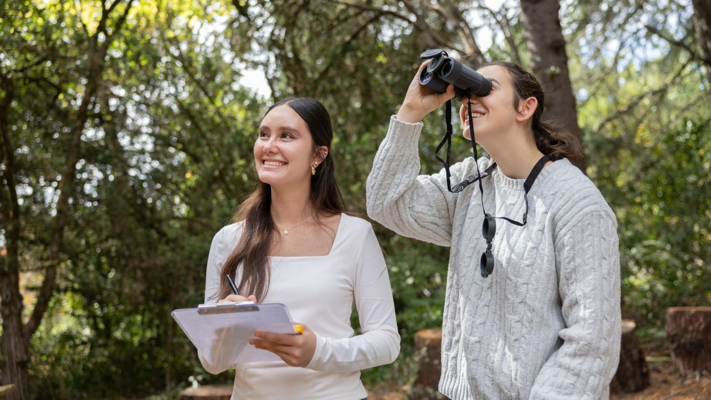 Two students outside with one holding a clipboard and one looking through binoculears